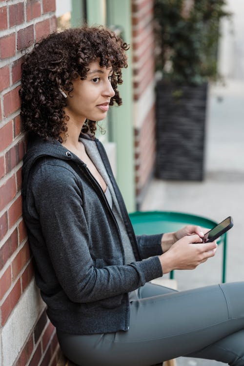 Young ethnic woman in TWS earphones using smartphone on bench in city ·  Free Stock Photo