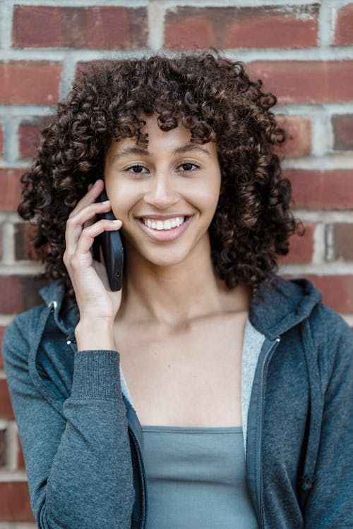 Cheerful young ethnic female with dark curly hair smiling and looking at camera while having phone conversation near brick wall on street