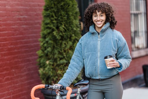 Content young ethnic woman with takeaway coffee smiling on street standing near bicycle