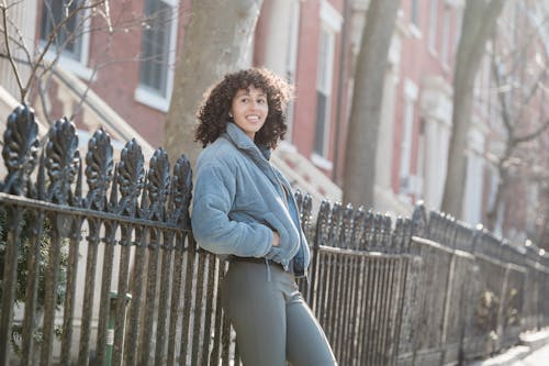 Side view of self assured young ethnic lady with curly hair in warm jacket leaning on metal fence and looking away while standing on city street on sunny day