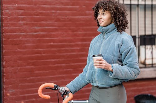 Free Content young ethnic lady with dark curly hair in warm walking with bicycle on street near brick building and drinking coffee to go Stock Photo