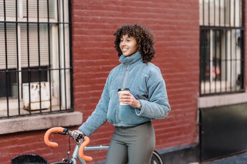 Free Happy young ethnic woman standing near bicycle on street and smiling while drinking takeaway coffee near brick building Stock Photo