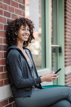 Side view of cheerful young female millennial with curly hair in activewear sitting on bench near brick building and smiling while talking on smartphone via TWS earphones