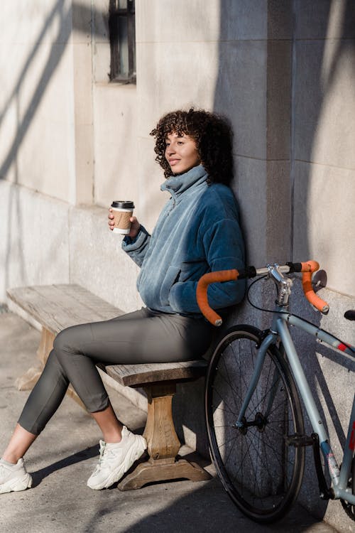 Free Side view of dreamy young ethnic woman with curly hair in casual clothes sitting on bench on street near parked bicycle and enjoying hot coffee in sunny morning Stock Photo