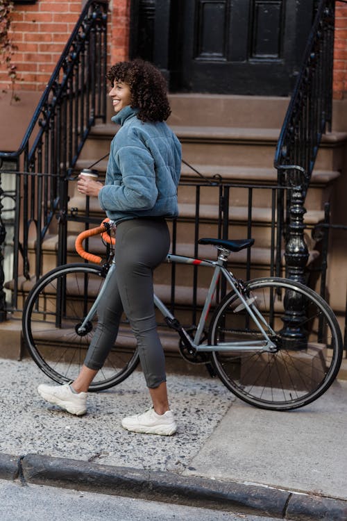 Free Side view of delighted young ethnic female with curly hair in warm clothes drinking coffee to go and smiling while walking on street with bicycle in city residential district Stock Photo