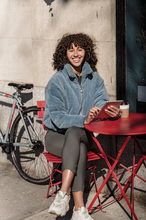 Cheerful ethnic female with cellphone and takeaway hot drink sitting with crossed legs at urban cafe table while looking at camera