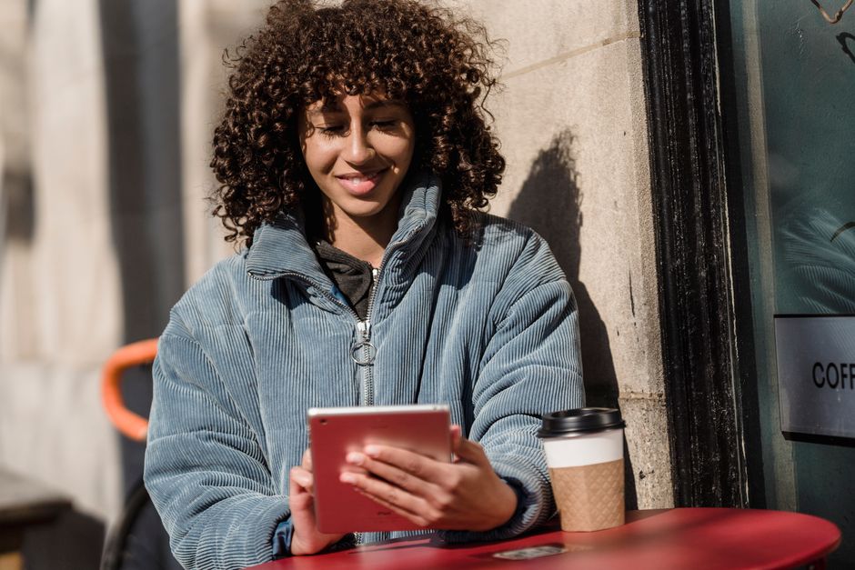 Cheerful ethnic female surfing internet on cellphone at table with hot beverage to go in street cafeteria on sunny day