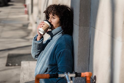 Side view of dreamy young ethnic female enjoying hot drink from paper cup while looking away in sunny town