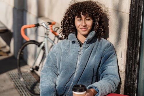 Contemplative ethnic female in jacket with hot drink to go looking away while sitting at urban cafe table in sunlight