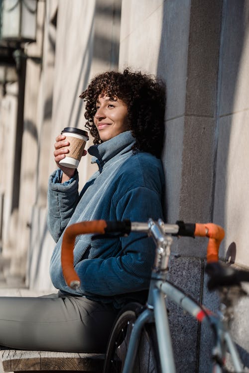 Side view of cheerful ethnic female with hand in pocket and hot drink to go looking away against bike in town