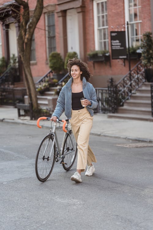 Smiling ethnic woman with coffee and bike walking on road
