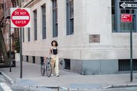 Contemplative ethnic woman with coffee and bike walking on pavement