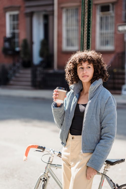 Free Calm young ethnic lady with dark curly hair in stylish outfit leaning on parked bicycle and drinking takeaway coffee on city street on sunny day Stock Photo