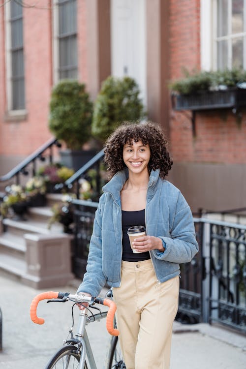Free Smiling young ethnic lady walking on street with bike and takeaway beverage Stock Photo