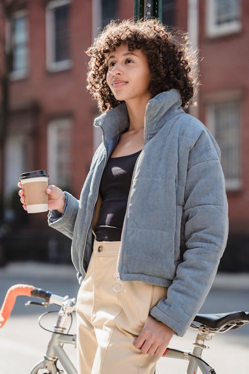 Free Dreamy young ethnic female millennial with curly hair in stylish warm outfit standing on street near parked bicycle and drinking coffee to go in sunny morning Stock Photo