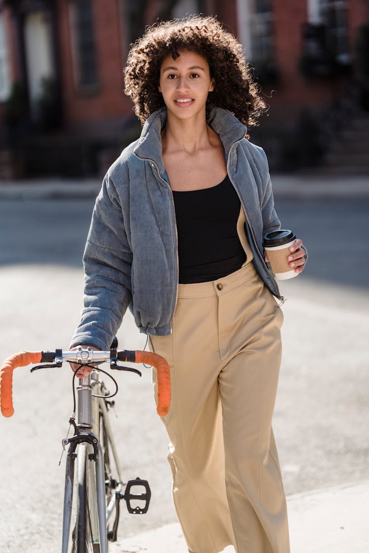 Young Ethnic Woman With Cup Of Takeaway Beverage Strolling On Street With Bike