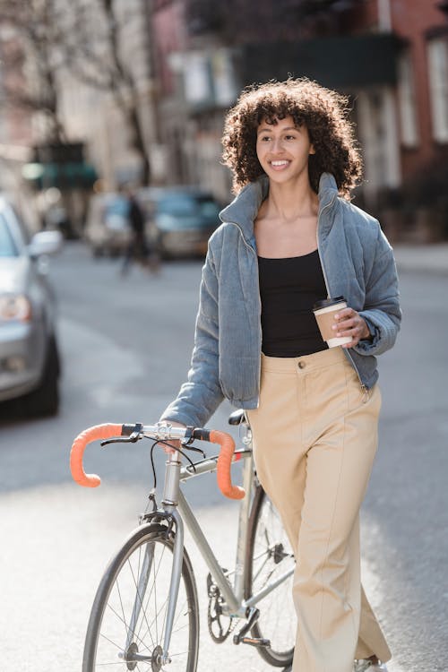 Cheerful young ethnic female with cup of takeaway coffee crossing road with bicycle