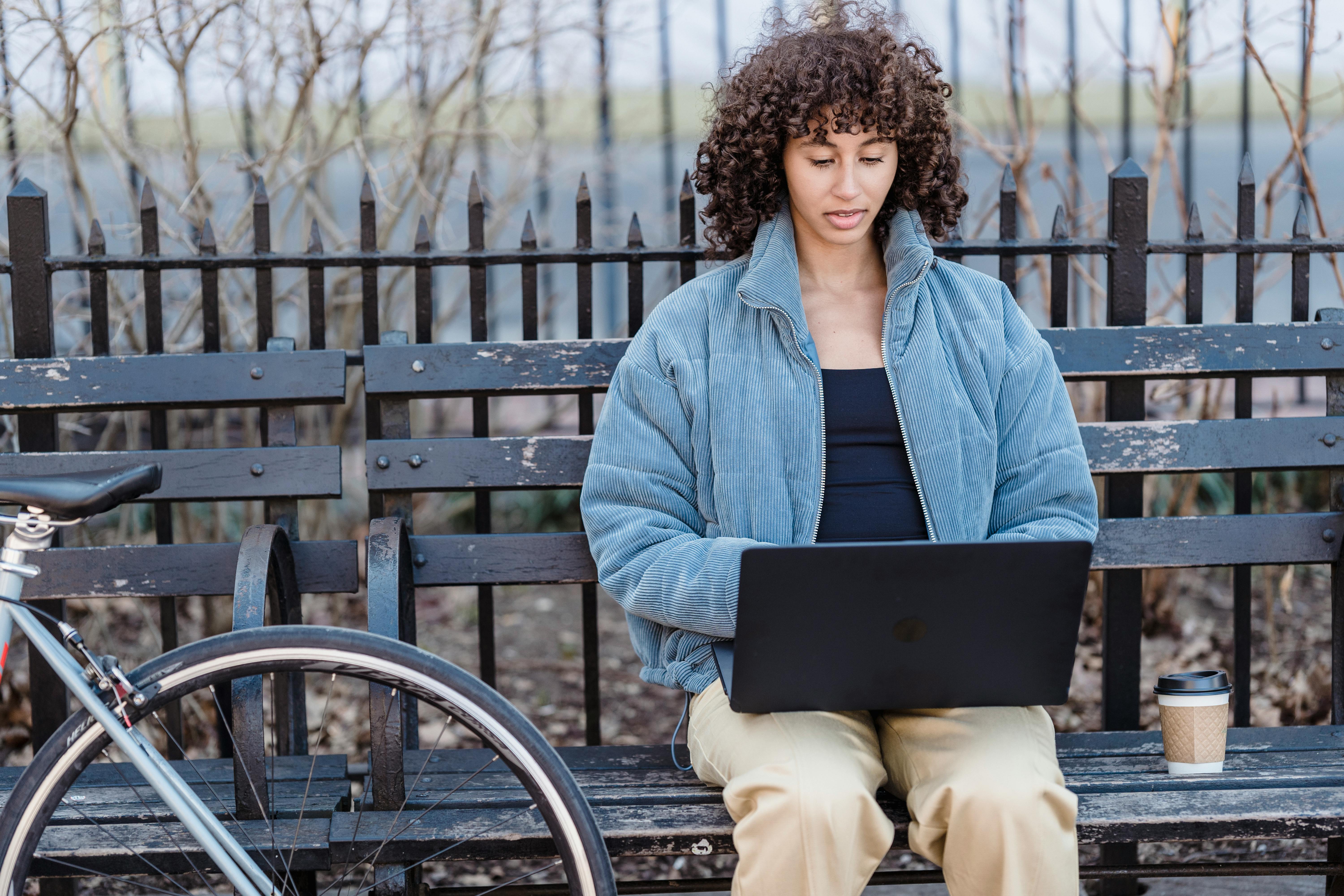 Young woman with curly hair working on a laptop outdoors in a park, signaling freelance lifestyle.