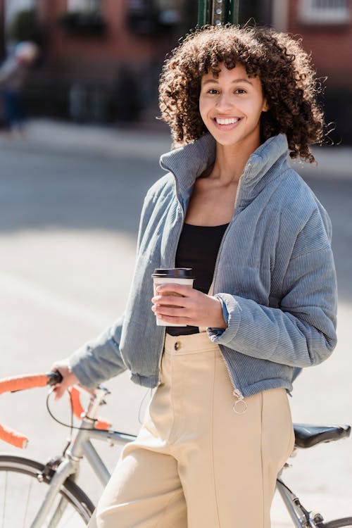 Free Joyful young ethnic female millennial with brown curly hair in casual warm outfit smiling happily and looking at camera while drinking takeaway coffee standing on street near parked bicycle Stock Photo