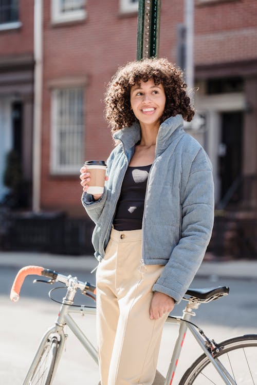 Free Happy young ethnic woman standing near bicycle on street and enjoying hot drink Stock Photo