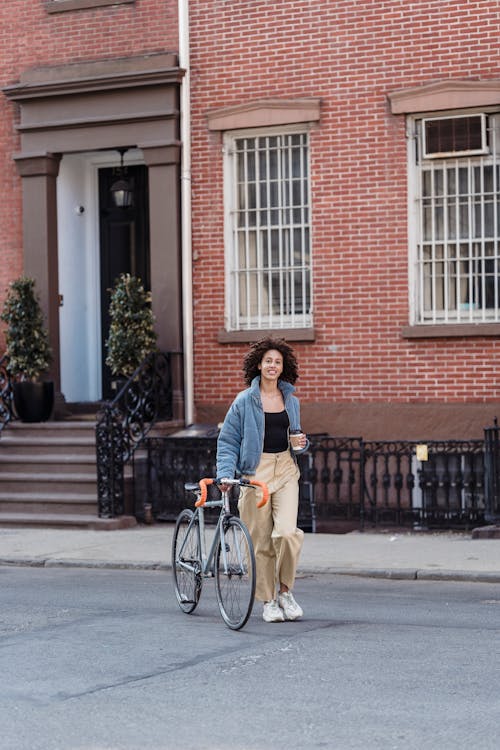 Full length of stylish young ethnic female cyclist rolling bicycle walking on city street near brick residential building