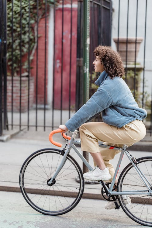 Side view of content young ethnic female millennial with curly hair in casual clothe smiling while riding bicycle on asphalt road in city residential district