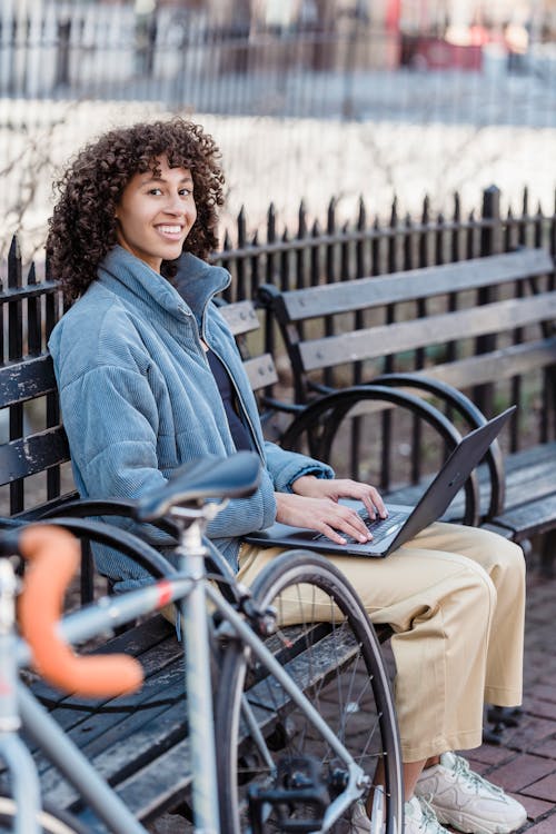 Free Positive young ethnic female freelancer with curly hair in trendy warm jacket smiling and looking at camera while browsing laptop sitting on wooden bench in city park Stock Photo