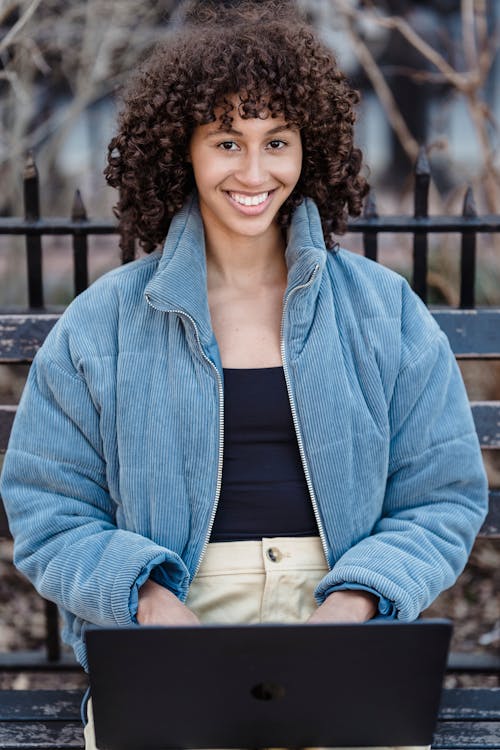 Confident young ethnic female freelancer with curly brown hair in casual outfit smiling and looking at camera while working distantly on laptop sitting on bench in autumn park