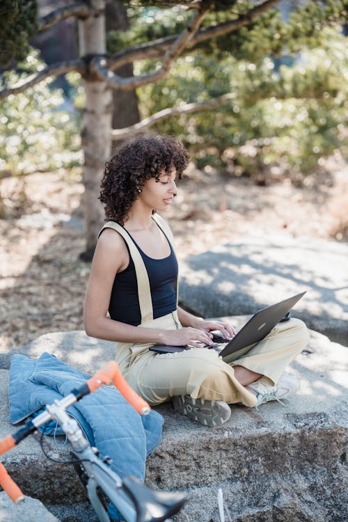 Free Side view of young ethnic female remote worker with curly hair in casual sitting on stone with crossed legs and working on laptop in green park Stock Photo