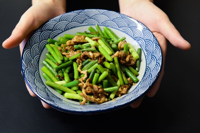 A delicious stir-fry of pork and green vegetables in a blue patterned bowl held by hands.