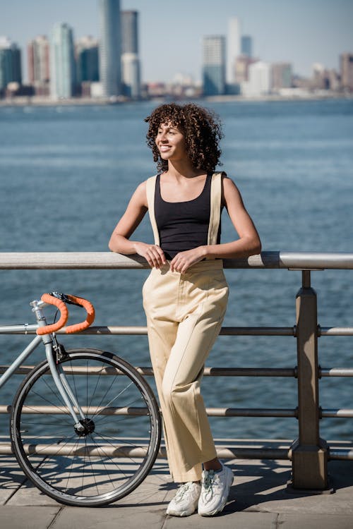 Full body of young female in trendy clothes standing with bicycle on seafront near river in city on sunny day