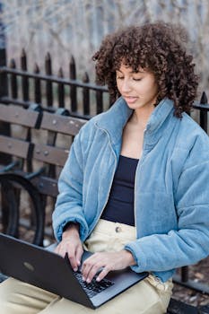 A young woman with curly hair types on a laptop while sitting on a park bench outdoors.