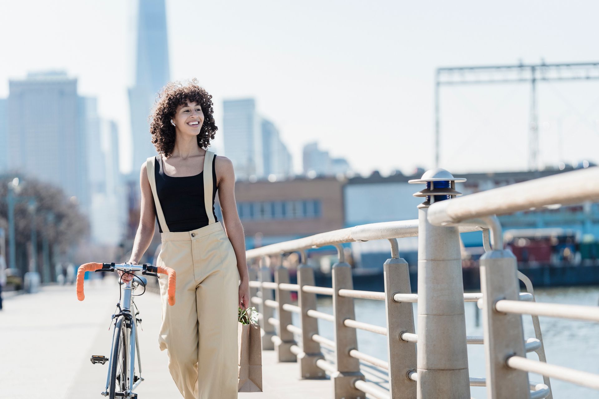 Young ethnic female in casual clothes walking with bike on city seafront and smiling while looking away