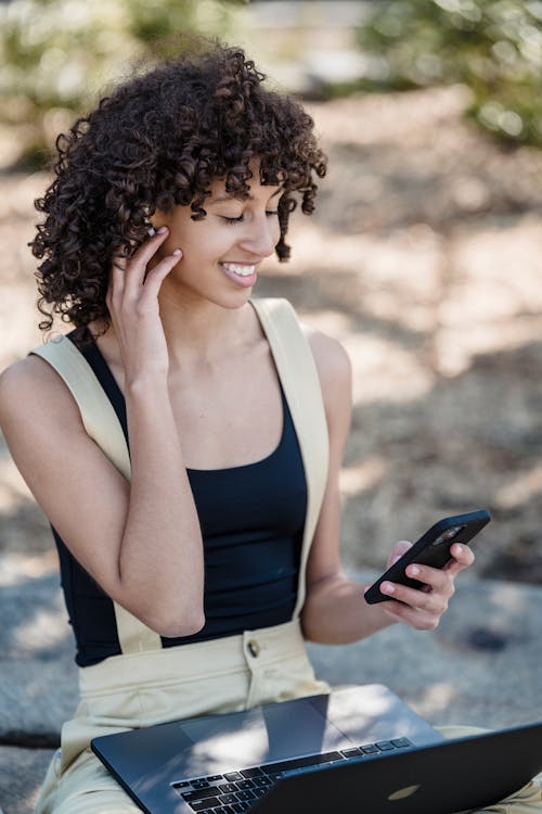 Young ethnic female in casual clothes with Afro hairstyle browsing phone while sitting with laptop on stone in park