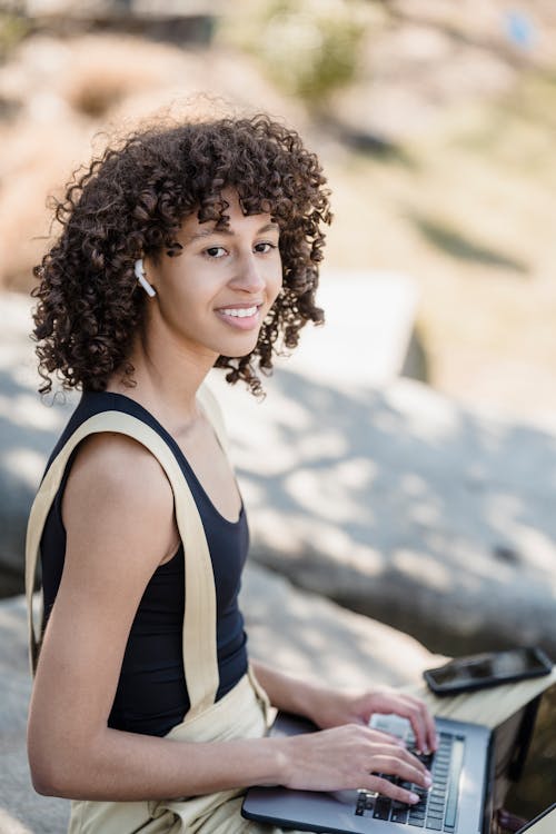 Side view of young ethnic female with Afro hairstyle in casual clothes sitting on stone border and looking away while browsing laptop