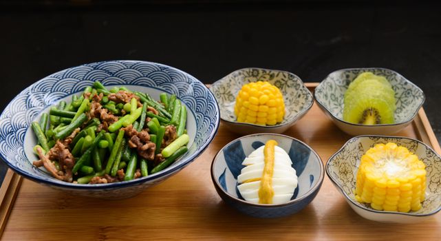 A vibrant food arrangement featuring a healthy mix of vegetables, corn, egg, and kiwi on a wooden tray.