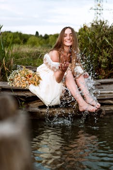 An elegant woman sits by a river, playfully splashing water, surrounded by nature. by Kiro Wang