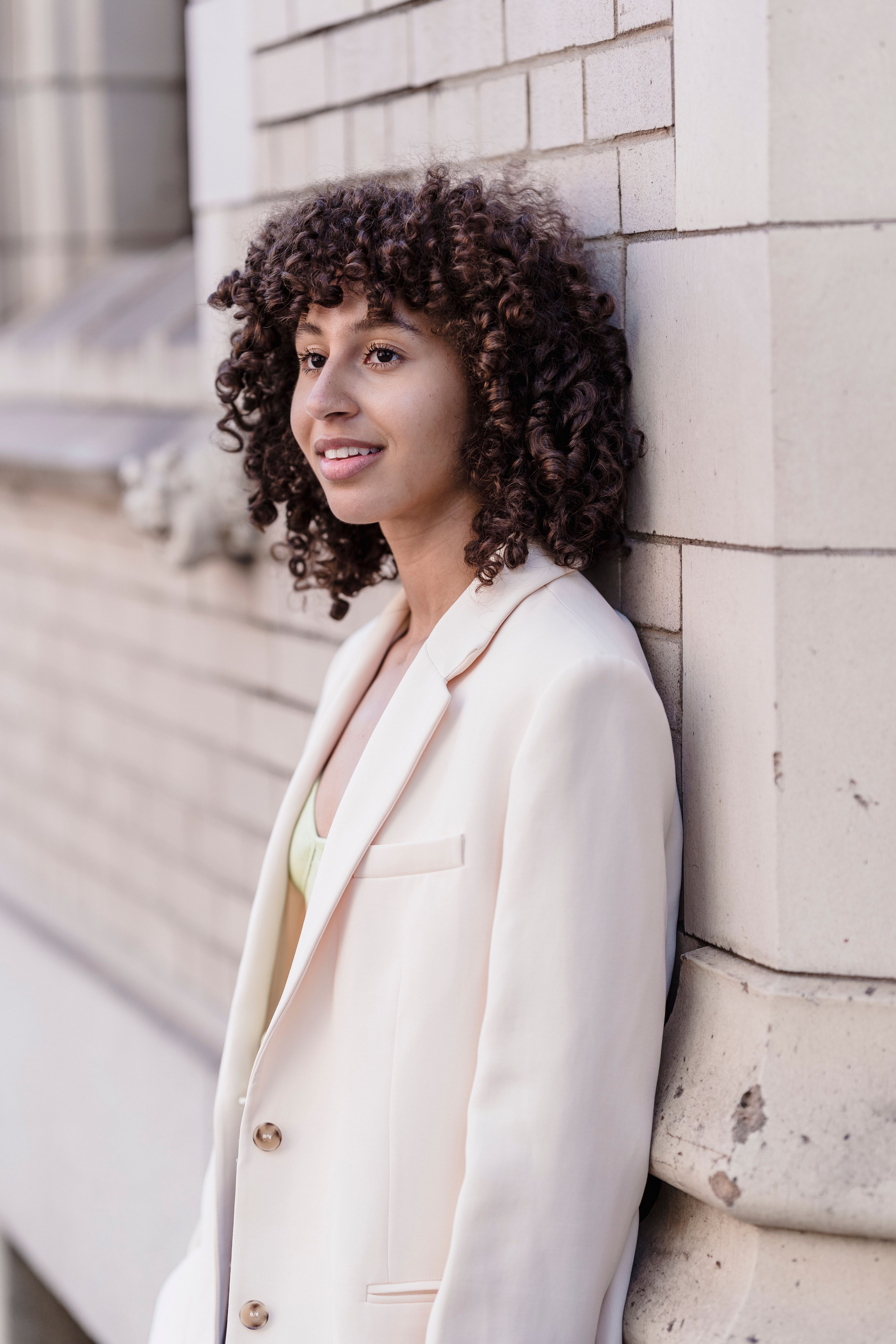 Portrait of a smiling woman with curly hair leaning on a brick wall, wearing a white blazer.