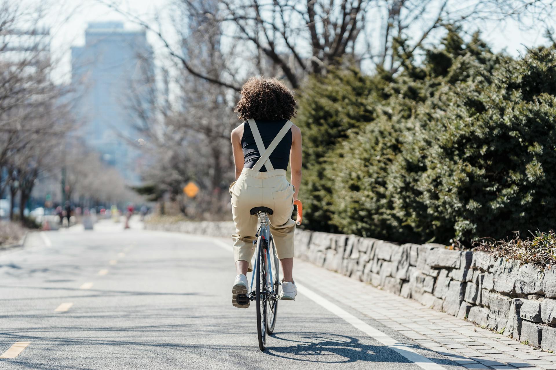 Back view of black anonymous female cyclist riding bicycle on asphalt road in city with green lush bushes aside