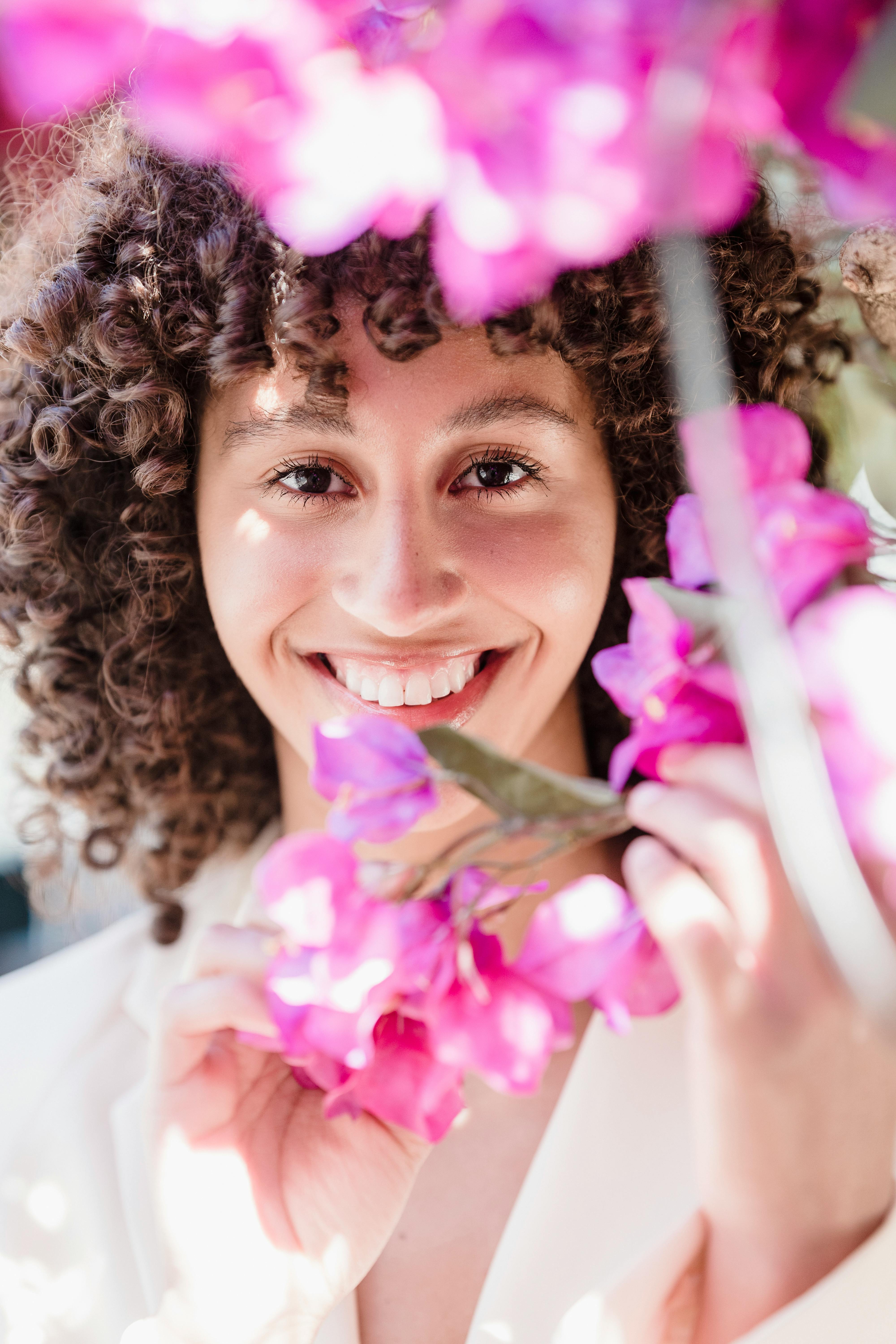 Portrait of satisfied black woman holding tender pink flowers against face and looking at camera