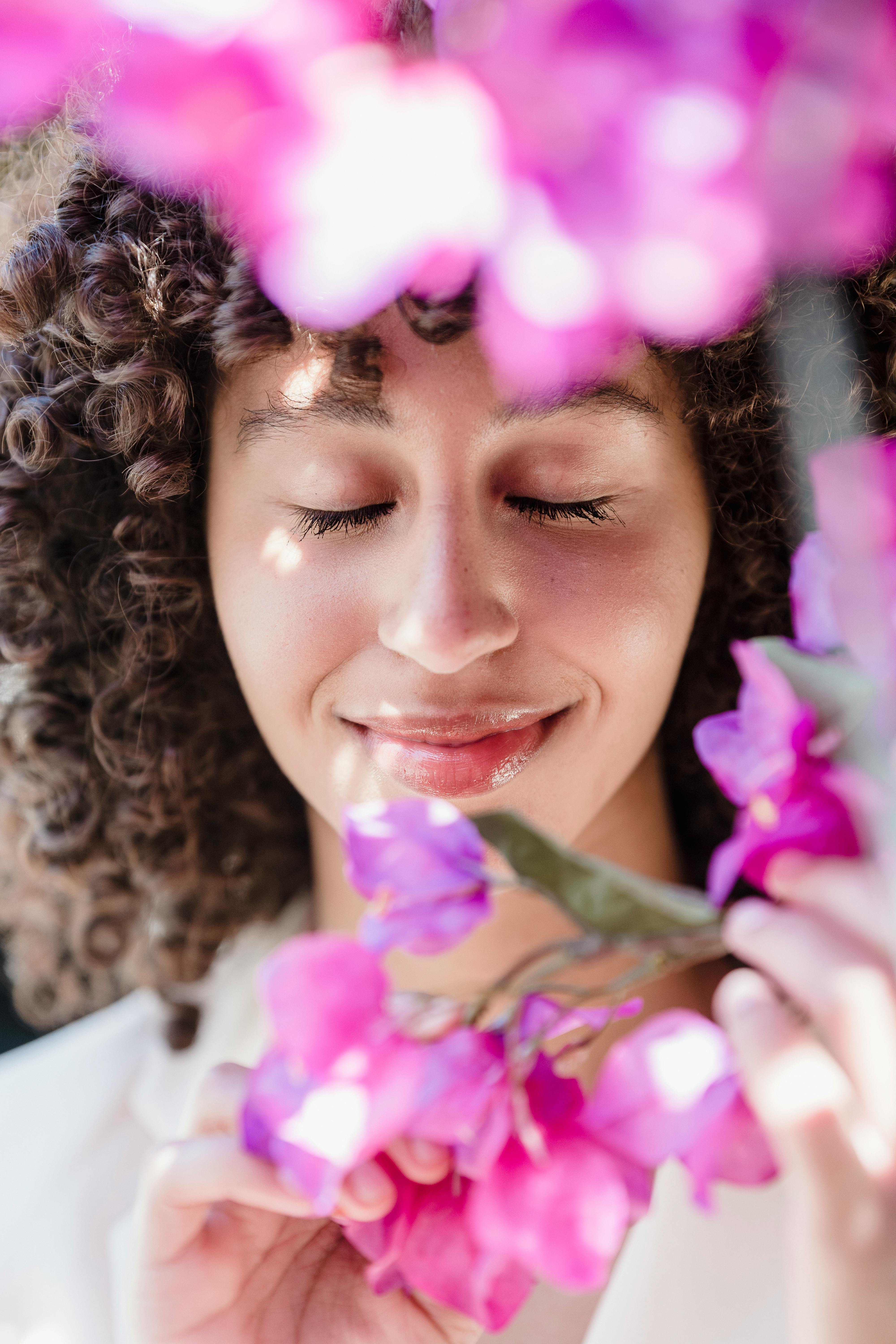 Content young ethnic lady with dark curly hair smiling with closed eyes while enjoying smell of fresh blooming flowers on sunny day in garden