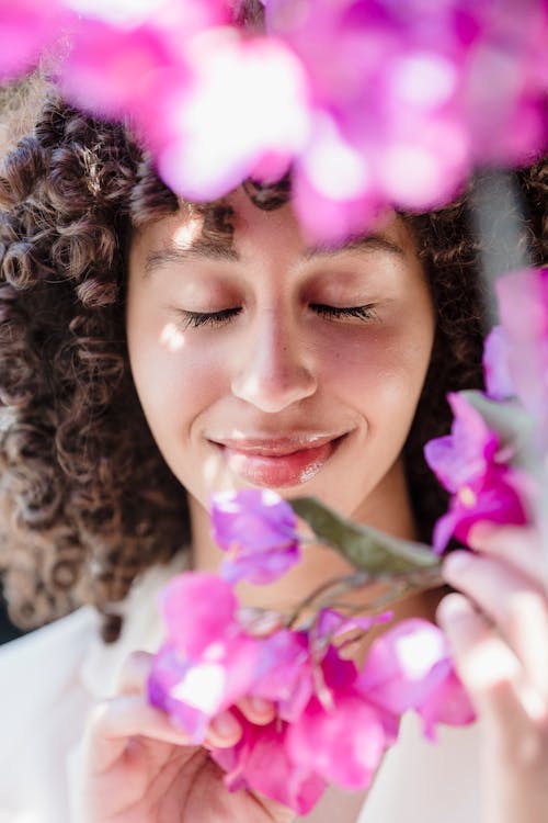 Content young ethnic lady with dark curly hair smiling with closed eyes while enjoying smell of fresh blooming flowers on sunny day in garden