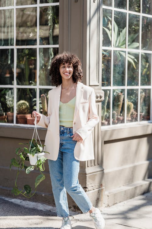 Cheerful ethnic female with green potted plant in beige jacket smiling on sunny urban street