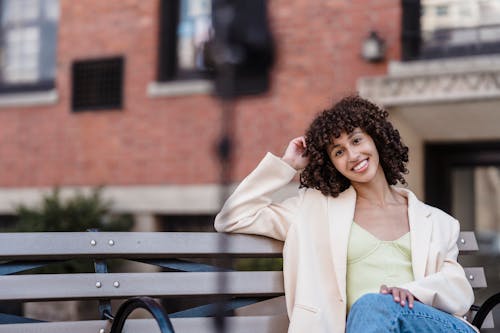 Young positive ethnic female touching curly dark hair smiling and looking at camera on blurred background of urban building