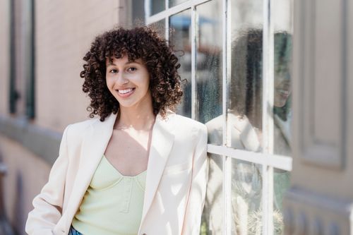Free Cheerful ethnic young female smiling and looking at camera while leaning on window of house Stock Photo