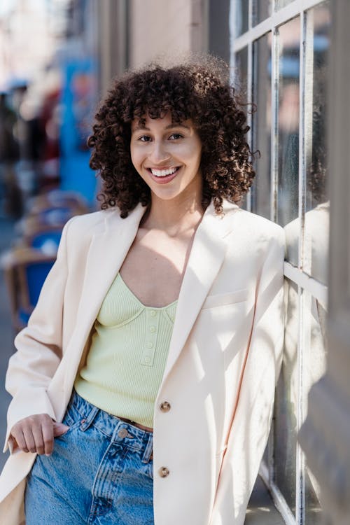 Cheerful fashionable ethnic female with curly hair smiling and looking at camera while leaning on window of building
