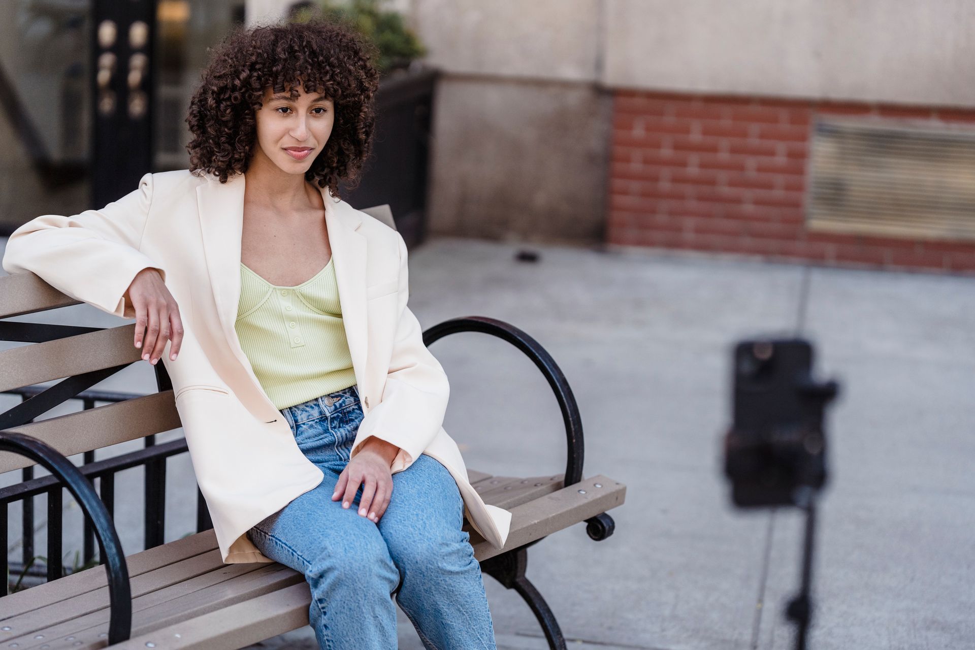 Young ethnic female in white elegant jacket and jeans resting on bench while talking online on mobile phone