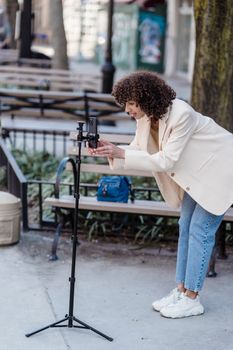 Side view of young ethnic female vlogger leaning forward to cellphone on tripod before video record in town