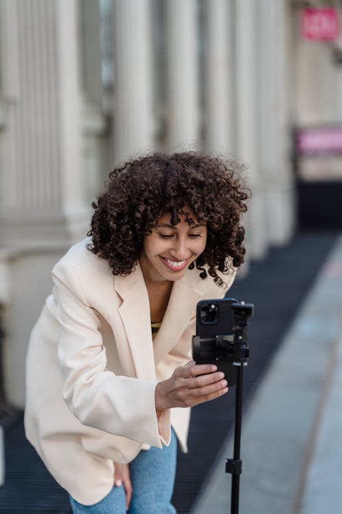 Content ethnic female vlogger preparing cellphone for video record while leaning forward on pavement in daytime
