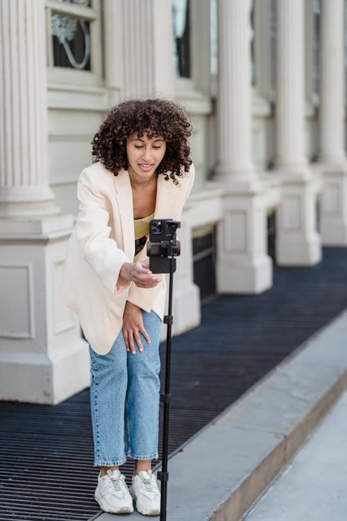 Free Young ethnic female vlogger with curly hair leaning forward to cellphone on tripod before video record on walkway Stock Photo